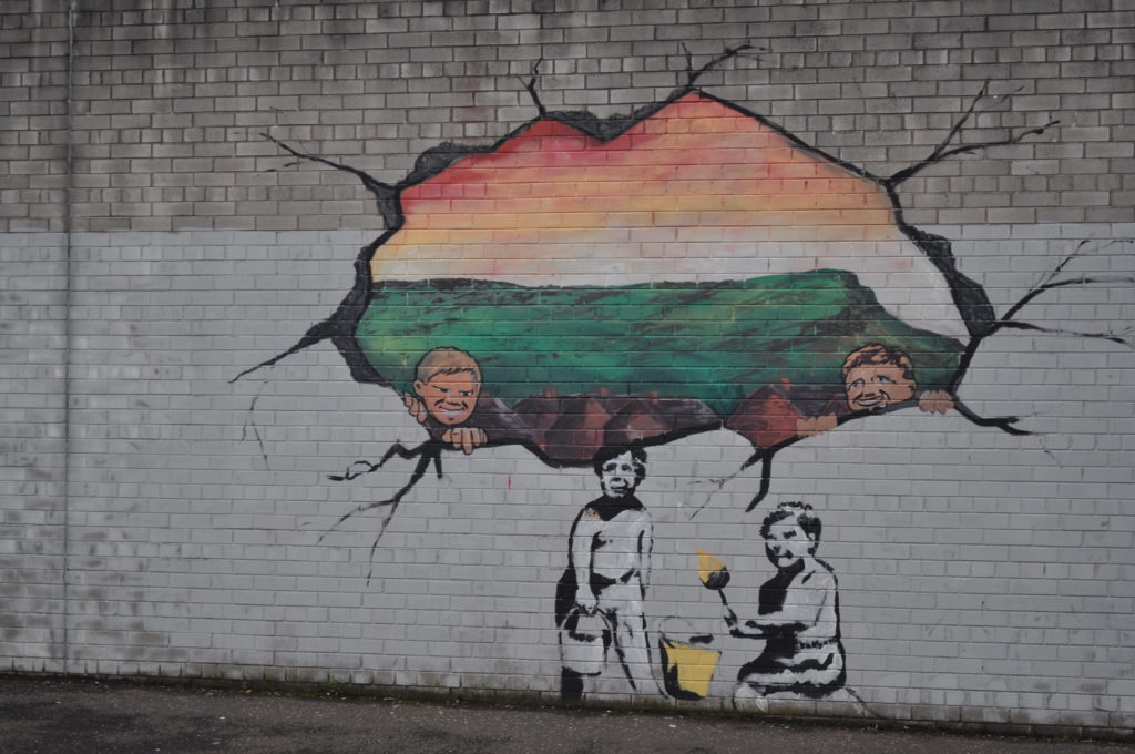 Il murale (di Gabriele Scalet) sulla Falls Road mostra l'Irlanda simboleggiata in un paesaggio idilliaco che sfugge attraverso le fessure dell'Irlanda del Nord